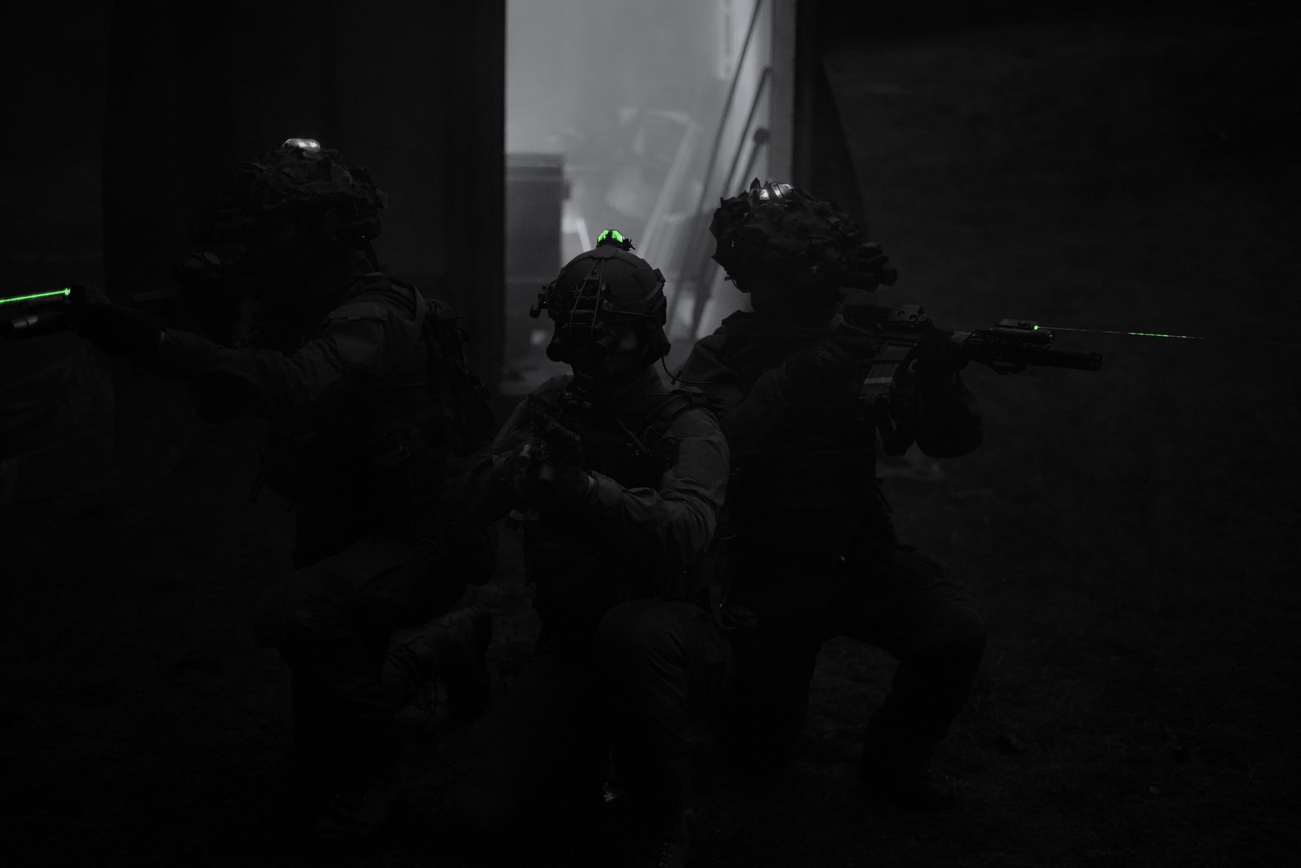 Three soldiers wearing night vision devices kneel in the dark outside a building.