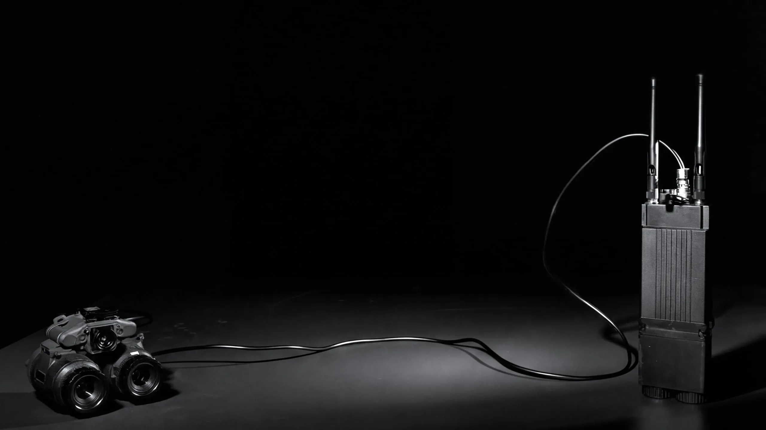 A night vision binocular on a dark floor, cabled to a radio with two antennas.